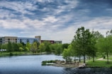 A scenic view of a lakeside park with lush green trees and a calm body of water. A modern building complex is visible in the background under a partly cloudy sky. The park features walking paths, rocks, and well-maintained grass.