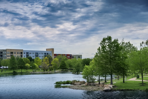 A scenic view of a lakeside park with lush green trees and a calm body of water. A modern building complex is visible in the background under a partly cloudy sky. The park features walking paths, rocks, and well-maintained grass.