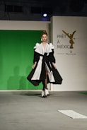 A model walks down a runway wearing a striking black and white gown with voluminous sleeves and a high slit. The backdrop features a green screen with a logo for 'Prêt à México' and a reference to Maserati. The atmosphere suggests a fashion show setting with professional lighting.