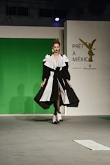 A model walks down a runway wearing a striking black and white gown with voluminous sleeves and a high slit. The backdrop features a green screen with a logo for 'Prêt à México' and a reference to Maserati. The atmosphere suggests a fashion show setting with professional lighting.