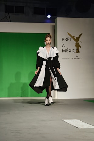 A model walks down a runway wearing a striking black and white gown with voluminous sleeves and a high slit. The backdrop features a green screen with a logo for 'Prêt à México' and a reference to Maserati. The atmosphere suggests a fashion show setting with professional lighting.