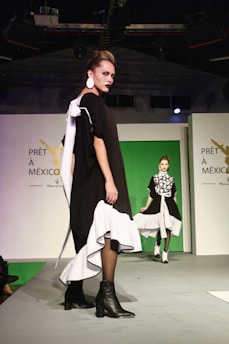 A fashion runway scene features two models wearing stylish black and white outfits with distinctive designs. The foreground model has a black dress with bold white accents and is wearing black ankle boots. Her hair is styled upwards, and she wears striking makeup. The background model wears a similar color-schemed outfit with a patterned top. The stage backdrop displays the words 'PRÊT À MEXICO.'