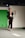 A model walks on the runway wearing a fashionable black outfit with feathered accents on the sleeves and shoulders. The outfit consists of a cropped top and high-waisted pants paired with black high heels. The backdrop includes a green panel and a white wall with a logo featuring an emblematic figure.
