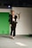 A model walks on the runway wearing a fashionable black outfit with feathered accents on the sleeves and shoulders. The outfit consists of a cropped top and high-waisted pants paired with black high heels. The backdrop includes a green panel and a white wall with a logo featuring an emblematic figure.
