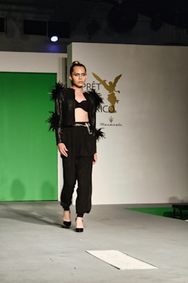 A model walks on the runway wearing a fashionable black outfit with feathered accents on the sleeves and shoulders. The outfit consists of a cropped top and high-waisted pants paired with black high heels. The backdrop includes a green panel and a white wall with a logo featuring an emblematic figure.