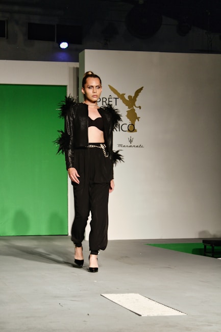 A model walks on the runway wearing a fashionable black outfit with feathered accents on the sleeves and shoulders. The outfit consists of a cropped top and high-waisted pants paired with black high heels. The backdrop includes a green panel and a white wall with a logo featuring an emblematic figure.