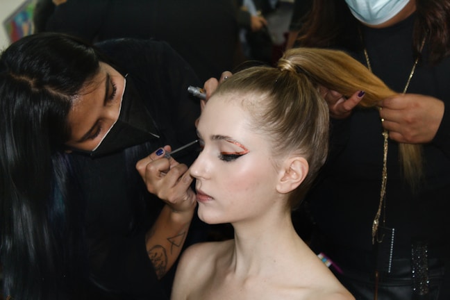A makeup artist applies eyeliner to a young woman with her hair pulled back. Another person holds the woman's ponytail. The focus is on the detailed eye makeup, showing skillful artistry.