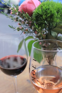 A close-up view of two glasses of wine, one red and one rosé, set on a table with a bouquet of flowers in the background consisting of a pink rose and green foliage.