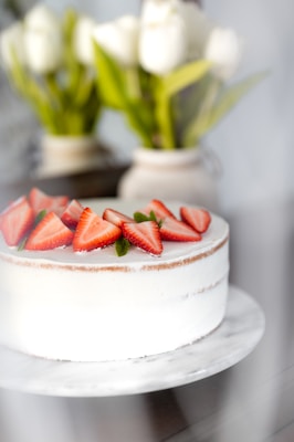 A beautifully decorated cake with a smooth white frosting and fresh strawberry slices arranged on top. The cake sits on a marble platter. In the background, there are blurred white flowers in vases, adding an elegant touch to the scene.