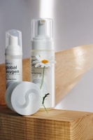 Two skincare products with pump dispensers and a round container are placed on a wooden surface. A white daisy flower with a yellow center is positioned next to the larger bottle, adding a natural element to the composition. The labels display branding and text, with a minimalistic design featuring dots and curves.