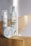 Two skincare products with pump dispensers and a round container are placed on a wooden surface. A white daisy flower with a yellow center is positioned next to the larger bottle, adding a natural element to the composition. The labels display branding and text, with a minimalistic design featuring dots and curves.
