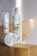 Two skincare products with pump dispensers and a round container are placed on a wooden surface. A white daisy flower with a yellow center is positioned next to the larger bottle, adding a natural element to the composition. The labels display branding and text, with a minimalistic design featuring dots and curves.