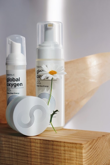 Two skincare products with pump dispensers and a round container are placed on a wooden surface. A white daisy flower with a yellow center is positioned next to the larger bottle, adding a natural element to the composition. The labels display branding and text, with a minimalistic design featuring dots and curves.