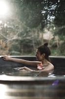 A person with their hair tied up is relaxing in a circular outdoor hot tub. Sunlight filters through surrounding trees, creating a tranquil and serene atmosphere. The background is blurred, emphasizing the calmness and natural setting.