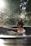 A person with their hair tied up is relaxing in a circular outdoor hot tub. Sunlight filters through surrounding trees, creating a tranquil and serene atmosphere. The background is blurred, emphasizing the calmness and natural setting.