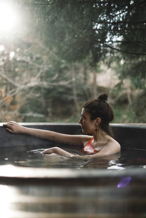 A person with their hair tied up is relaxing in a circular outdoor hot tub. Sunlight filters through surrounding trees, creating a tranquil and serene atmosphere. The background is blurred, emphasizing the calmness and natural setting.