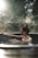 A person with their hair tied up is relaxing in a circular outdoor hot tub. Sunlight filters through surrounding trees, creating a tranquil and serene atmosphere. The background is blurred, emphasizing the calmness and natural setting.