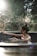 A person with their hair tied up is relaxing in a circular outdoor hot tub. Sunlight filters through surrounding trees, creating a tranquil and serene atmosphere. The background is blurred, emphasizing the calmness and natural setting.