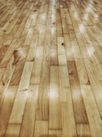 A detailed view of a wooden floor with parallel planks neatly arranged. The surface shows natural wood grain patterns with varying shades. There are some spots and slight imperfections visible on the wood, adding character to the surface.