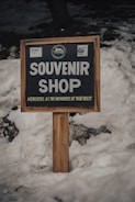 A wooden sign with bold white letters reads 'Souvenir Shop' and 'Mementos as the memories of your visit' underneath. It is set against a backdrop of snow with several rocks partially visible at the bottom left corner.