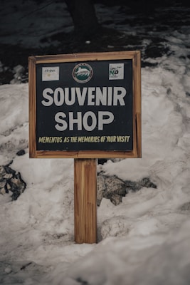 A wooden sign with bold white letters reads 'Souvenir Shop' and 'Mementos as the memories of your visit' underneath. It is set against a backdrop of snow with several rocks partially visible at the bottom left corner.