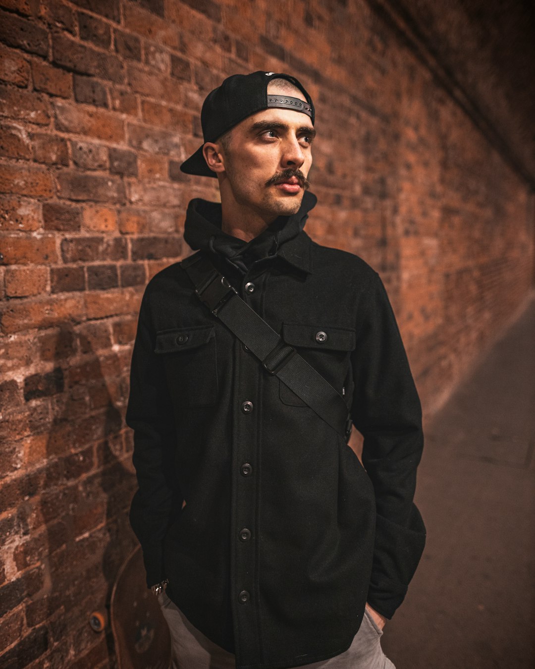 man in black button up long sleeve shirt and black cap standing beside brown brick wall