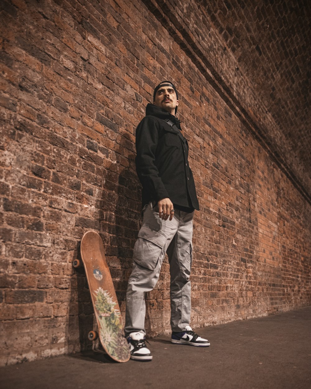 man in black jacket and white pants standing beside brown brick wall