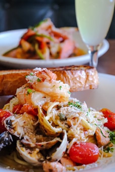 A plate of seafood pasta garnished with grated cheese, fresh herbs, and accompanied by a slice of toasted bread. The dish includes shrimp, clams, cherry tomatoes, and a creamy sauce. In the background, there is another plate with a similar dish and a tall glass of a light-colored beverage.