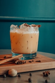 A glass filled with a creamy, iced beverage garnished with cinnamon sticks and star anise, sitting on a wooden board. Scattered around the board are whole spices including nutmeg and cloves. The background is a muted teal wall, contributing to a rustic and cozy atmosphere.