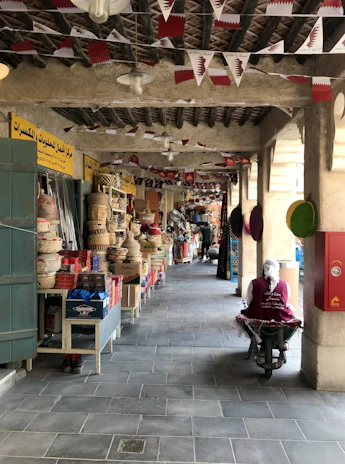 A market corridor adorned with maroon and white flags featuring national symbols. The pathway is lined with various stalls selling woven baskets, colorful textiles, and other traditional goods. A figure wearing traditional attire sits on a cart towards the right, while warm lighting illuminates the scene.