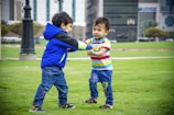 Two young children are playing together on a grassy field. One wears a colorful striped shirt and blue jeans, while the other is dressed in a bright blue jacket and denim pants. Both appear engaged and joyful in their interactions, set against an urban backdrop with a tall lamp post and buildings.