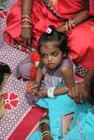 A young girl with a small flower hair accessory sits surrounded by adults wearing vibrant traditional clothing. She has dark eye makeup, and is sitting on a patterned fabric with colorful designs. The adults around her are wearing ornate bangles and richly colored sarees.