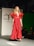 A model in a striking red gown with ruffled sleeves walks down a runway. The garment is floor-length and features a cinched waistline. The background includes a backdrop with text indicating 'PRÊT À MEXICO' and a seated audience observing the fashion show.
