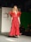 A model in a striking red gown with ruffled sleeves walks down a runway. The garment is floor-length and features a cinched waistline. The background includes a backdrop with text indicating 'PRÊT À MEXICO' and a seated audience observing the fashion show.
