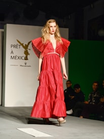 A model in a striking red gown with ruffled sleeves walks down a runway. The garment is floor-length and features a cinched waistline. The background includes a backdrop with text indicating 'PRÊT À MEXICO' and a seated audience observing the fashion show.