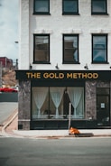 A storefront named 'The Gold Method' featuring a white facade with black-framed windows and a sign with gold lettering. The large windows display sheer white curtains and the lower portions have text advertising services. A street corner with a curb and an orange traffic cone is visible in front of the building.