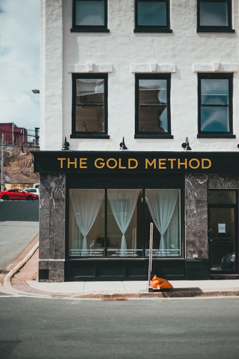 A storefront named 'The Gold Method' featuring a white facade with black-framed windows and a sign with gold lettering. The large windows display sheer white curtains and the lower portions have text advertising services. A street corner with a curb and an orange traffic cone is visible in front of the building.