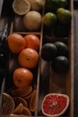 An assortment of citrus fruits is arranged in wooden compartments. There are whole oranges and several lime-colored fruits. Sliced sections of grapefruit with vibrant red pulp and several cut orange wedges are also visible.