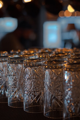 A collection of intricately patterned crystal glasses is arranged closely together on a dark surface. The glasses reflect and refract ambient light, creating subtle highlights and shadows in the dimly lit environment. The background is blurred, featuring warm, out-of-focus lights that contribute to the overall ambiance.