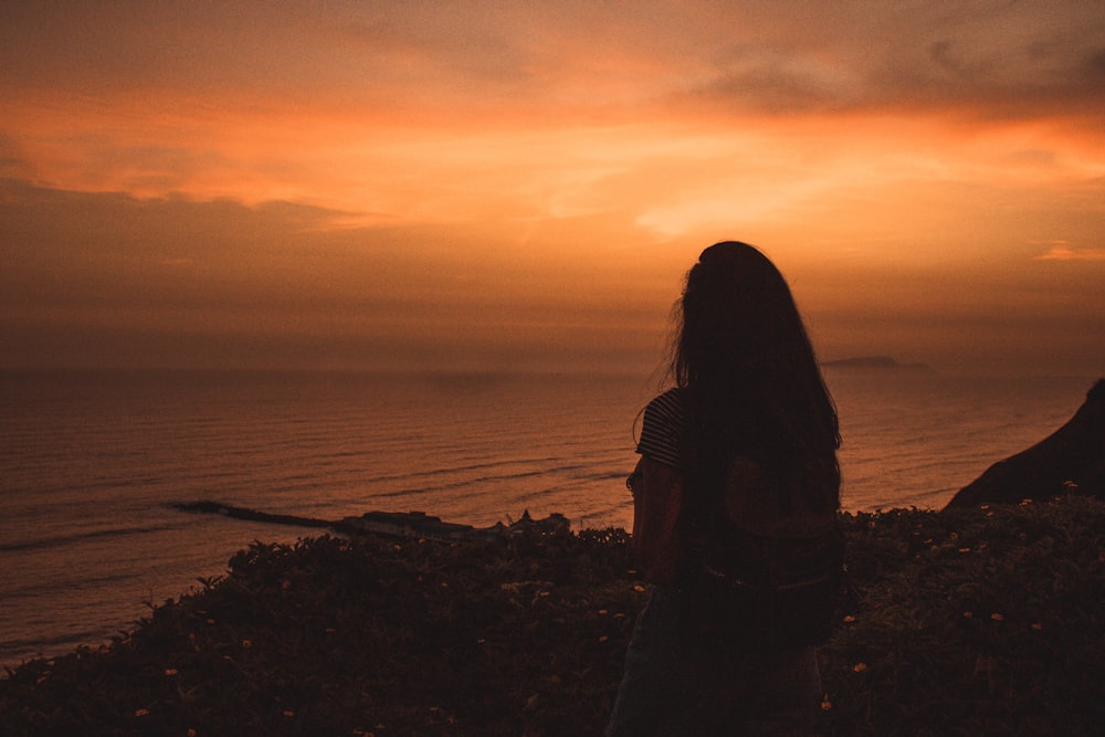 silhouette of woman standing near body of water during sunset