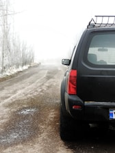 A black SUV is parked on a foggy, rural road. The ground appears wet and muddy, with patches of ice or snow visible. Leafless trees line the sides of the road, disappearing into the dense fog in the distance. The license plate on the SUV indicates it's from Iran.