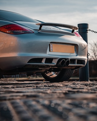 A sleek silver sports car is parked on a paved surface, with its back end prominently displayed. The car features a rear spoiler, dual exhaust pipes, and distinctive red tail lights. A pole with a chain can be seen next to the car, suggesting a parking area or pathway.