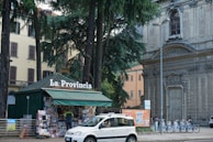 A small kiosk with a green roof is surrounded by tall trees. The kiosk displays a sign, 'La Provincia', and is filled with newspapers and magazines. A person stands at the counter. Nearby, a white car is parked on the street. Behind the kiosk, there is a historic stone building with intricate architectural details, including a large arched window and ornate carvings, and another building with yellow walls.