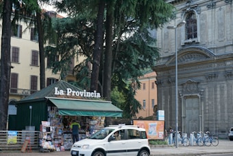 A small kiosk with a green roof is surrounded by tall trees. The kiosk displays a sign, 'La Provincia', and is filled with newspapers and magazines. A person stands at the counter. Nearby, a white car is parked on the street. Behind the kiosk, there is a historic stone building with intricate architectural details, including a large arched window and ornate carvings, and another building with yellow walls.