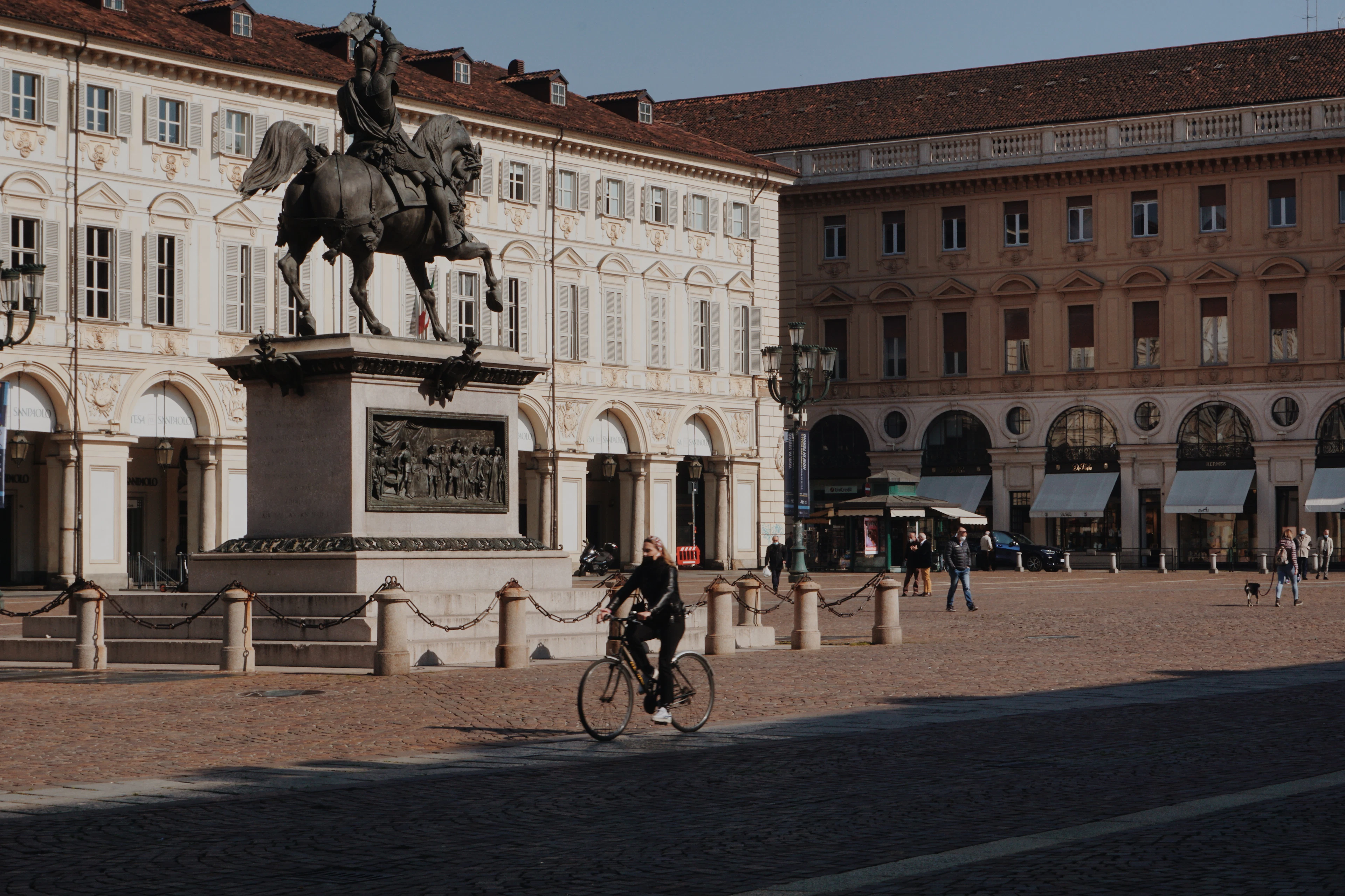A cyclist crossing the wide cobbled expanse of Piazza San Carlo in Turin in low afternoon sun, passing the bronze equestrian statue with the square's arcaded white and ochre palazzi behind.