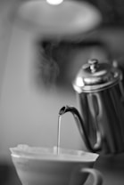 A close-up view of a metal kettle pouring hot water into a filter placed over a cup. Steam is visibly rising from the water, adding a sense of warmth and freshness to the image. The background is blurred, focusing attention on the kettle and cup.