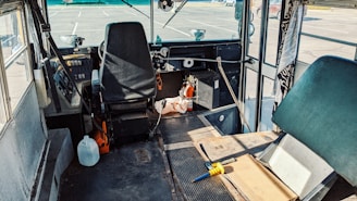The interior of a bus or van with a driver's seat and steering wheel, surrounded by various items such as a plastic jug, tools, and cleaning supplies. The windshield and controls are visible, with a somewhat cluttered space around the driver's area. Sunlight is casting shadows across the dashboard and floor.