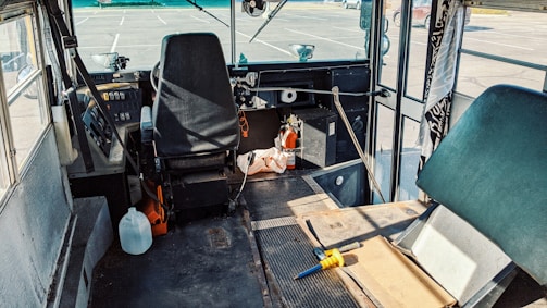The interior of a bus or van with a driver's seat and steering wheel, surrounded by various items such as a plastic jug, tools, and cleaning supplies. The windshield and controls are visible, with a somewhat cluttered space around the driver's area. Sunlight is casting shadows across the dashboard and floor.