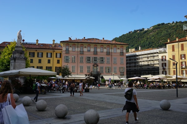 A lively town square with people walking and sitting at outdoor cafes. Surrounding the square are colorful historic buildings with balconies. A statue is visible on the left, and there is a backdrop of green hills under a clear blue sky.
