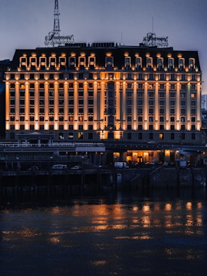 A large, elegant hotel building with multiple windows is illuminated by warm lights, creating reflections on the water in front. The structure's façade features classical architectural elements, and an ornate sign is visible on the rooftop. The scene is set during the evening, capturing the serene ambiance of the area.
